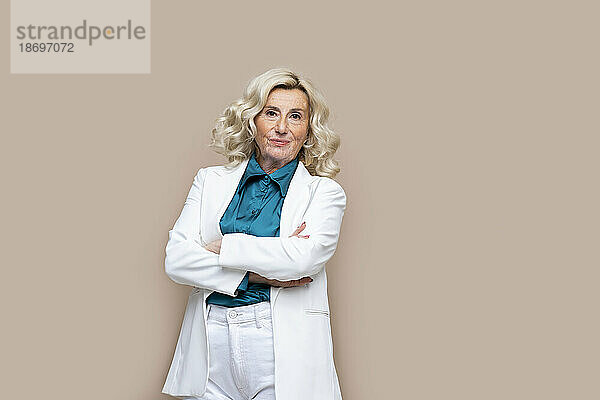 Smiling blond senior businesswoman with arms crossed against beige background