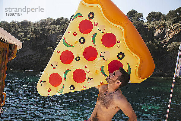 Mann ohne Hemd hält im Urlaub eine Pizza-Luftmatratze in der Hand