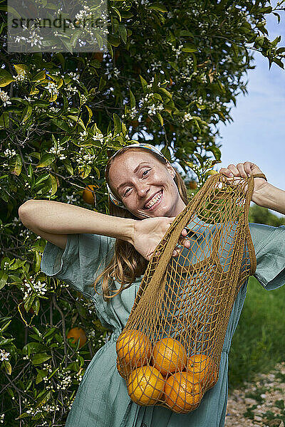 Fröhliche Frau mit frischen Orangen im Netzbeutel im Obstgarten