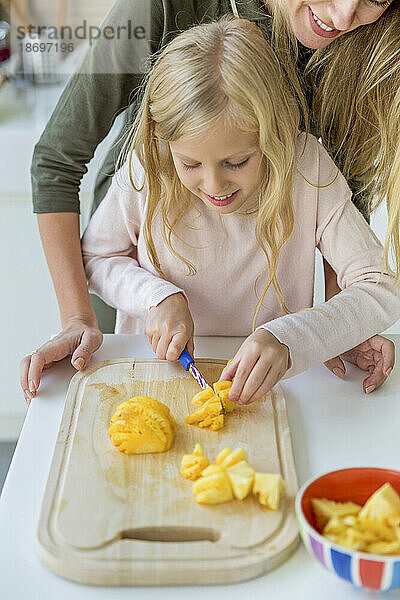 Lächelndes Mädchen lernt von der Mutter zu Hause  Ananas auf einem Schneidebrett zu hacken