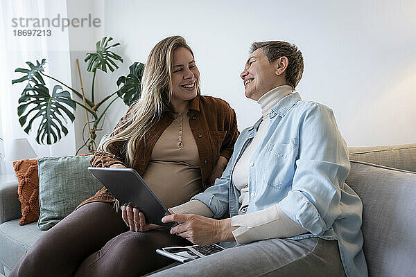 Glückliche Mutter mit Tablet-PC und schwangere Tochter  die zu Hause auf dem Sofa sitzt
