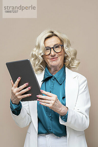 Lächelnde ältere Geschäftsfrau hält Tablet-PC vor beigem Hintergrund