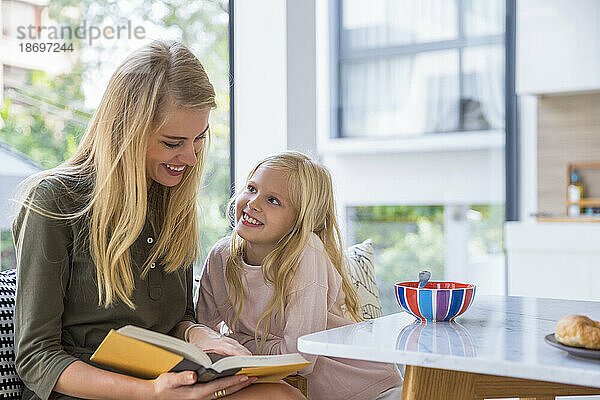 Lächelndes Mädchen schaut Mutter zu  wie sie zu Hause in der Küche ein Buch liest