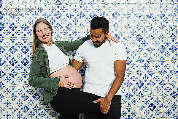 Glücklicher Mann mit schwangerer Frau vor gemusterter Fliesenwand