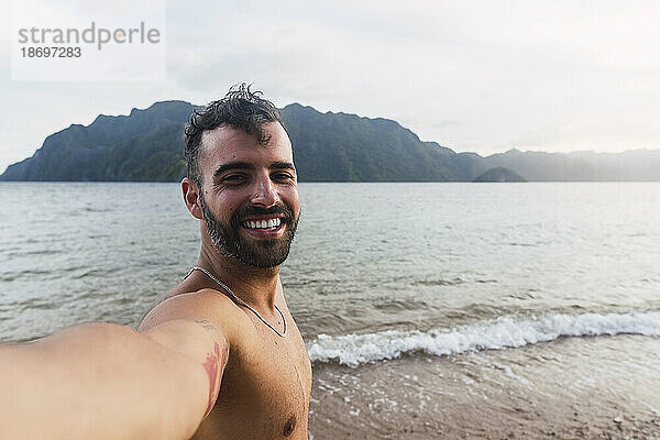 Glücklicher Mann ohne Hemd  der am Ufer ein Selfie macht