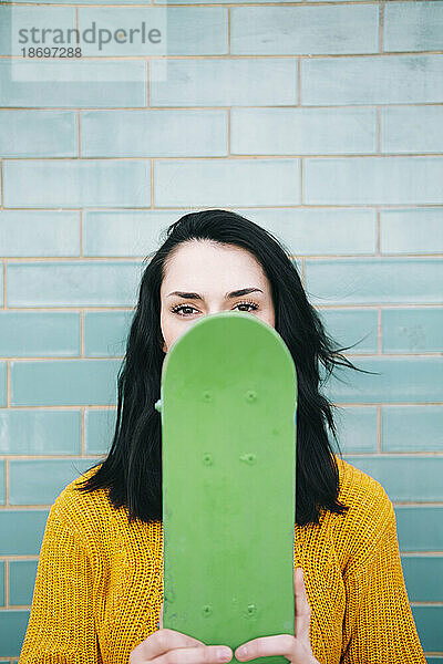 Junge Frau bedeckt Gesicht mit grünem Skateboard vor der Wand