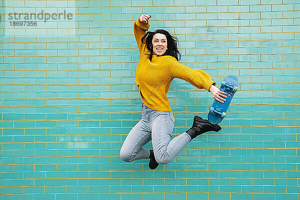 Fröhliche Frau hält Skateboard und springt hoch vor die Wand