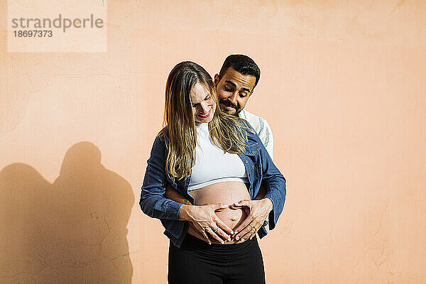 Smiling man making heart shape of pregnant woman's belly in front of wall