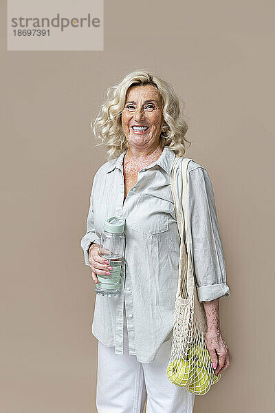 Smiling senior businesswoman with holding water bottle and mesh bag with fruits against beige background