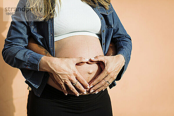 Schwangere Frau macht Herzform auf Bauch mit Mann vor Wand