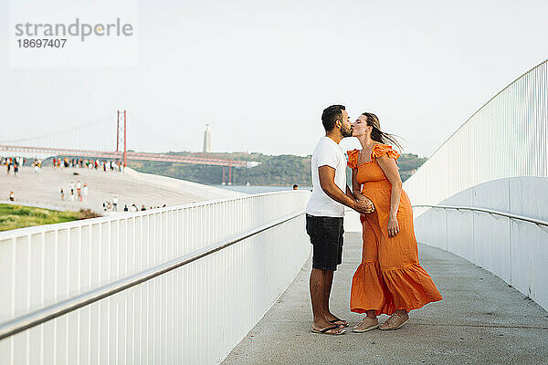 Schwangere Frau küsst Mann  der neben Geländer auf Fußweg steht