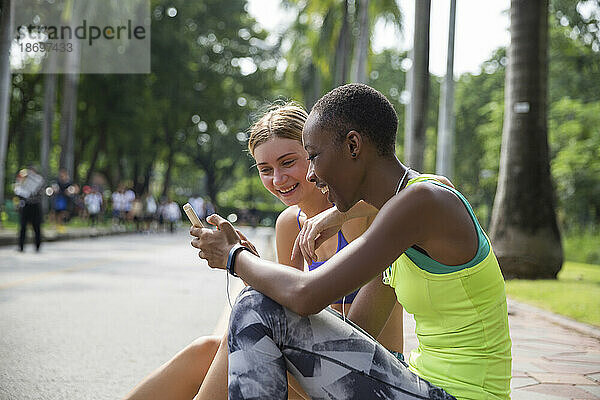 Glückliche Frau  die ihr Smartphone benutzt und mit einer Freundin auf dem Bürgersteig im Park sitzt
