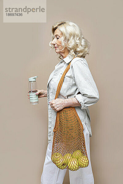 Senior businesswoman with mesh bag and water bottle against beige background