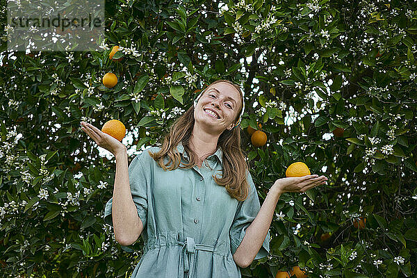 Lächelnde Frau hält Orangen neben Baum im Obstgarten