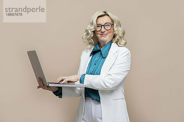 Lächelnde ältere Geschäftsfrau hält Laptop vor farbigem Hintergrund