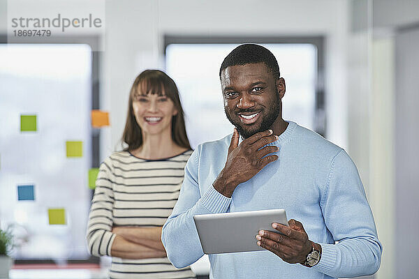 Lächelnder Geschäftsmann mit der Hand am Kinn  der einen Tablet-PC im Büro hält