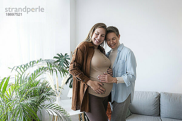 Lächelnde Mutter berührt den Bauch ihrer schwangeren Tochter  die zu Hause steht