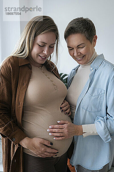 Smiling senior woman touching belly of pregnant daughter at home