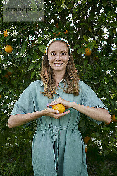 Lächelnde Frau mit frischer Orange vor dem Baum im Obstgarten