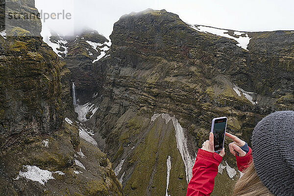 Nahaufnahme  von hinten aufgenommen  einer Frau  die steht und den Mulagljufur Canyon  ein Paradies für Wanderer  überblickt  während sie mit ihrem Smartphone ein Foto von einem atemberaubenden Blick auf einen Wasserfall durch die moosbedeckten Klippen macht; Vik  Südisland  Island