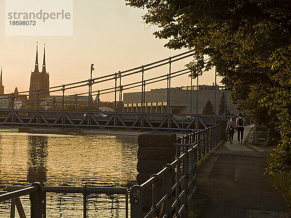 Paar spaziert bei Sonnenuntergang entlang der Oderpromenade mit Eisenbrücke und Kathedrale im Hintergrund; Breslau  Polen