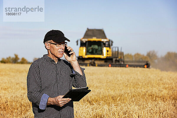 Landwirt nutzt ein Tablet  um seine Ernte zu verwalten  und telefoniert mit einem Mähdrescher im Hintergrund; Alcomdale  Alberta  Kanada