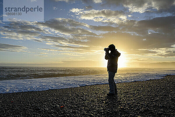 Silhouette eines Fotografen  der am Strand steht und bei Sonnenuntergang ein Foto von der Aussicht auf den Nordatlantik macht; Island