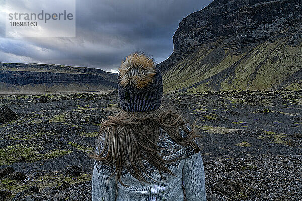 Nahaufnahme einer Frau mit blonden Haaren  aufgenommen von hinten  mit Wollmütze und Islandpullover  während sie in der Landschaft steht und die natürliche Schönheit der Berge und des stürmischen Himmels bewundert; Südisland  Island