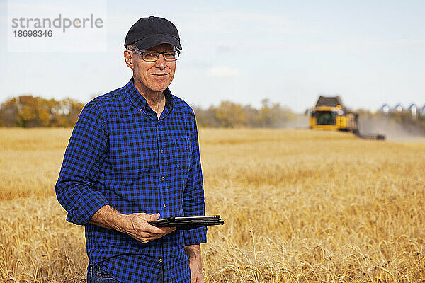 Landwirt blickt in die Kamera  während er mit einem Tablet seine Getreideernte verwaltet  während im Hintergrund Erntemaschinen arbeiten; Alcomdale  Alberta  Kanada