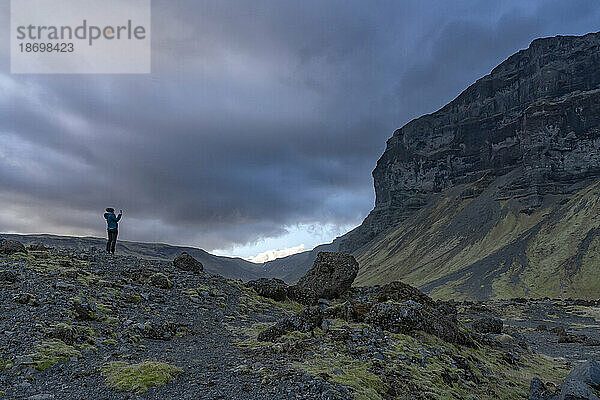 Eine Frau steht in der vulkanischen Felslandschaft  bewundert die natürliche Schönheit und fotografiert die Berge und die dramatischen Gewitterwolken  die sich über ihr bilden; Südisland  Island