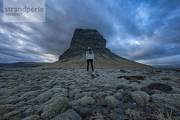Blick von hinten auf eine Frau  die eine Wollmütze und einen Islandpullover trägt  während sie in der Landschaft steht und die natürliche Schönheit der Berge und des stürmischen Himmels bewundert; Südisland  Island