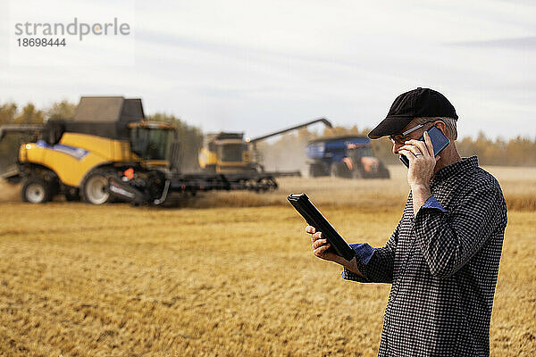 Landwirt nutzt ein Tablet  um seine Ernte zu verwalten  und telefoniert mit Erntemaschinen im Hintergrund; Alcomdale  Alberta  Kanada