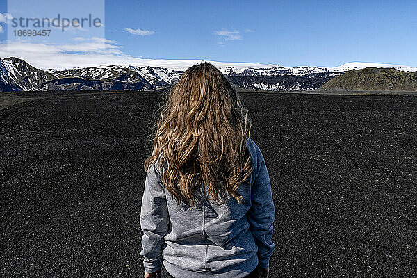 Nahaufnahme einer Frau mit blonden Haaren  aufgenommen von hinten  vor einer riesigen Landschaft aus schwarzem Sand mit einem Bergrücken und einem blauen Himmel in der Ferne; Island