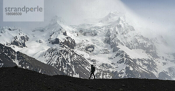Silhouette einer Person  die vor einem großen schneebedeckten Berg an der Südküste Islands entlang einer Gletschermoräne geht; Südisland  Island