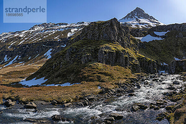 Nahaufnahme eines Flusses  der durch die atemberaubende Berglandschaft eines Fjords fließt  mit klarem  blauem Himmel; Ostisland  Island