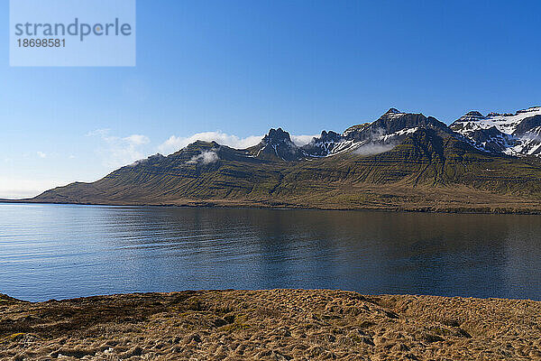 Das bergige Gelände der Ostfjorde mit blauem Himmel und ruhigem Wasser schafft eine atemberaubende Landschaft zum Durchreisen; Ostisland  Island