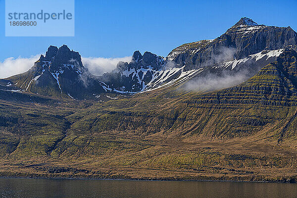 Nahaufnahme des bergigen Geländes der Ostfjorde mit blauem Himmel und nebligen Bergwolken  die eine atemberaubende Landschaft zum Durchreisen schaffen; Ostisland  Island