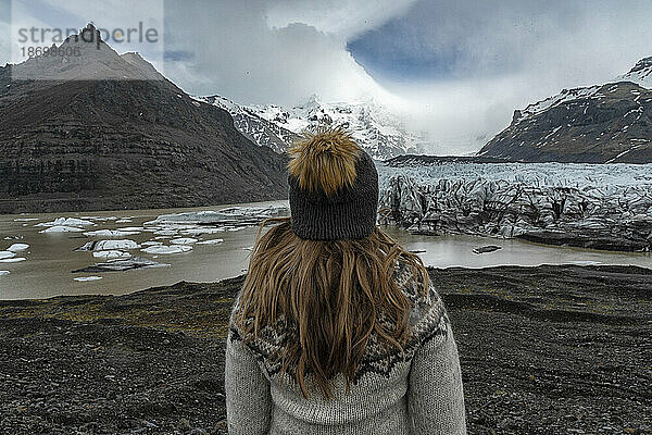 Nahaufnahme einer Frau mit blonden Haaren  aufgenommen von hinten  mit Wollmütze und Islandpullover mit Blick auf einen Gletscher und eine Lagune an der Südküste; Südisland  Island
