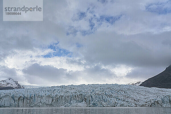 Blick von der Fjallsarlon-Gletscherlagune auf das blaue Eis der Endstation des Fjallsjökull-Gletschers  die sich von den grauen Wolken abhebt  die den blauen Himmel bedecken  am südlichen Ende des berühmten isländischen Gletschers Vatnajökull im Süden Islands; Südisland  Island