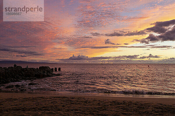 Blick vom Strand auf die Silhouette eines felsigen Piers  der ins Meer ragt  mit einem Himmel aus violetten und rosa Wolken und vor der Küste vertäuten Booten und dem goldenen Schein des Sonnenuntergangs  der sich im Wasser spiegelt; Nord-Kihei  Maui  Hawaii  Vereinigte Staaten von Amerika