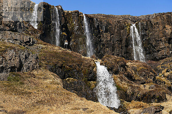 Szenische Ansicht einer Frau  die an einem Klippenhang vor einer Reihe von Wasserfällen steht  von denen einige oben und unten von den schroffen Klippen der Ostfjorde strömen und sie vor der weiten Landschaft vor ihr klein erscheinen lassen; Ostisland  Island