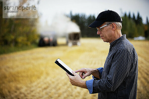 Landwirt nutzt ein Tablet  um seine Ernte zu verwalten  während im Hintergrund Erntemaschinen arbeiten; Alcomdale  Alberta  Kanada