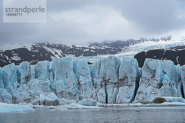 Der Blick auf das blaue Eis des Fjallsjökull-Gletschers hebt sich von den grauen  nebligen Wolken über den schneebedeckten Bergen ab  die man von der Gletscherlagune Fjallsarlon am südlichen Ende des berühmten isländischen Gletschers Vatnajökull im Süden Islands aus sieht. Südisland  Island