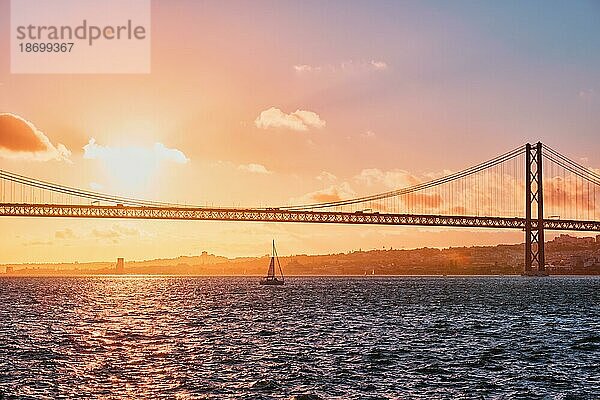 Blick auf die Brücke 25 de Abril  das berühmte touristische Wahrzeichen Lissabons  das Lisboa und Almada auf der Halbinsel Setubal verbindet  über den Fluss Tejo mit der Silhouette einer Touristenyacht bei Sonnenuntergang. Lissabon  Portugal  Europa