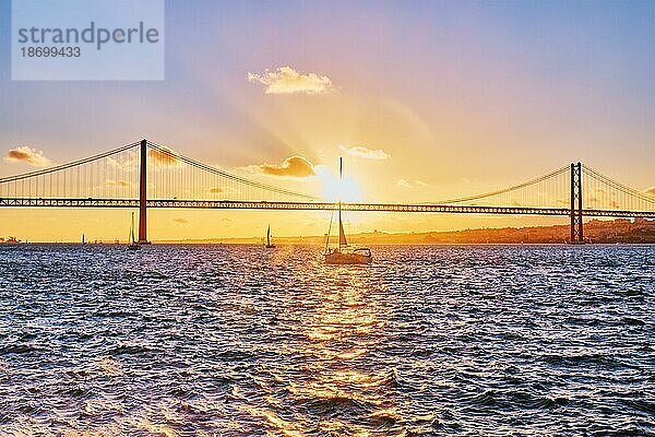 Blick auf die Brücke 25 de Abril  das berühmte touristische Wahrzeichen Lissabons  das Lisboa und Almada auf der Halbinsel Setubal verbindet  über den Fluss Tejo mit der Silhouette einer Touristenyacht bei Sonnenuntergang. Lissabon  Portugal  Europa