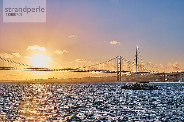 Blick auf die Brücke 25 de Abril  das berühmte touristische Wahrzeichen Lissabons  das Lisboa und Almada auf der Halbinsel Setubal verbindet  über den Fluss Tejo mit der Silhouette einer Touristenyacht bei Sonnenuntergang. Lissabon  Portugal  Europa