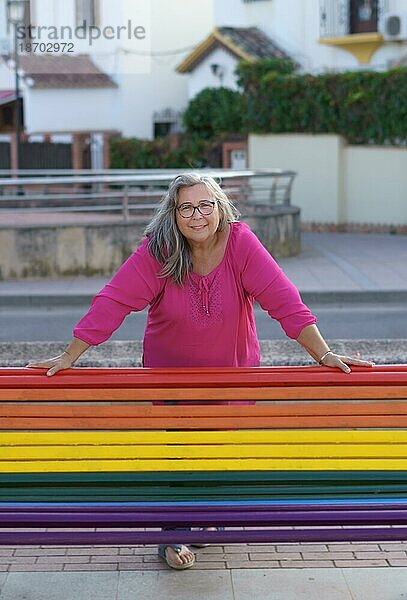 Ältere Frau mit weißen Haaren und rosa Hemd steht hinter einer Bank  die mit den Farben der LGTBIQ Flagge bemalt ist  und lächelt in die Kamera  queer