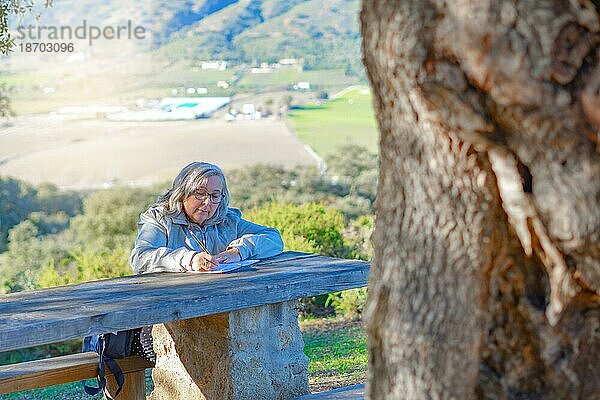 Frau sitzt auf einer Bank und schreibt in ein Notizbuch mit einer sonnenbeschienenen Berglandschaft im Hintergrund
