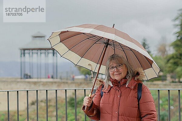 Weißhaarige Frau mit Mantel und Regenschirm an einem regnerischen Morgen schaut lächelnd in die Kamera