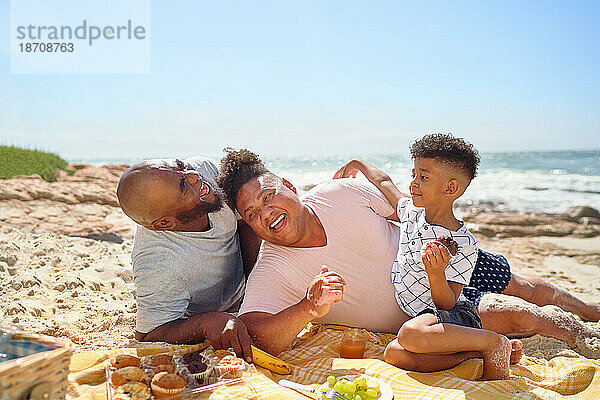 Glückliches schwules männliches Paar und Sohn  die am sonnigen Strand lachen und essen